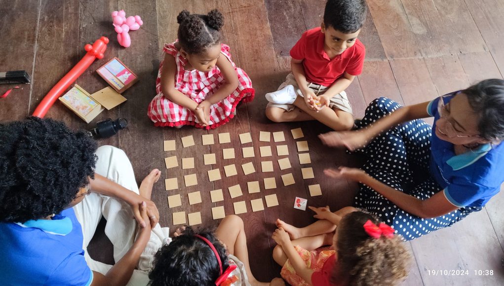 Foto de uma professora e crianças ao redor de um jogo da memória, aprendendo inglês.