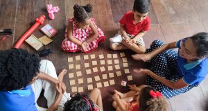 Aprender Inglês Desde Cedo: Benefícios para a Escola, a Carreira e a Vida Foto de uma professora e crianças ao redor de um jogo da memória, aprendendo inglês.