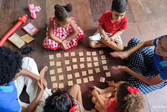 Aprender Inglês Desde Cedo: Benefícios para a Escola, a Carreira e a Vida Foto de uma professora e crianças ao redor de um jogo da memória, aprendendo inglês.
