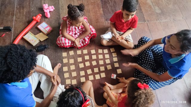 Foto de uma professora e crianças ao redor de um jogo da memória, aprendendo inglês.